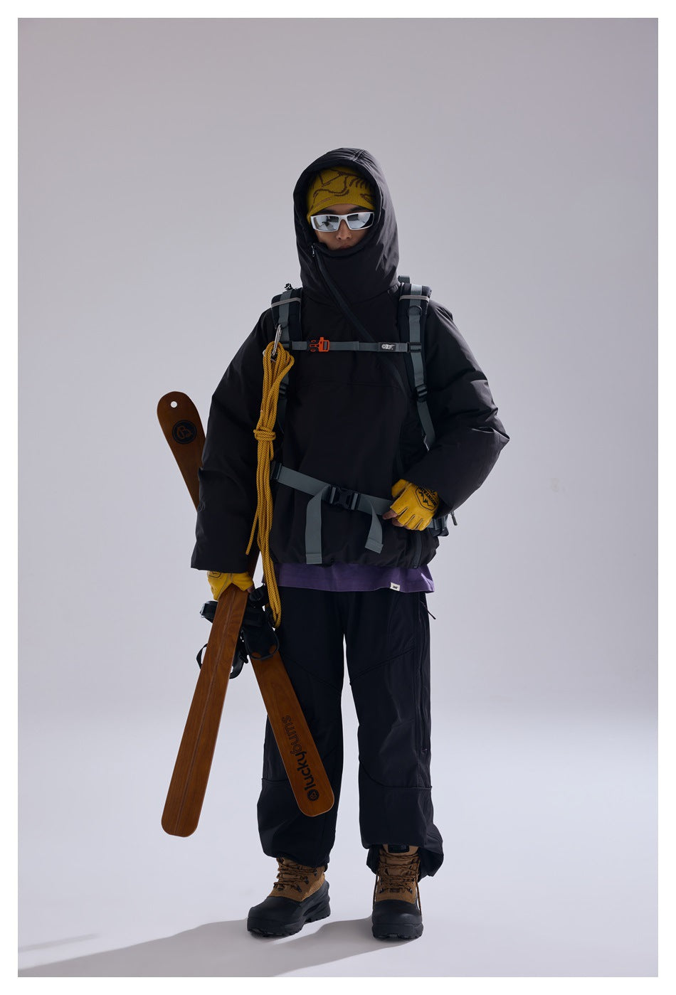 A person wearing a black hooded jacket and snow pants with a backpack harness, holding skis against a plain gray background.