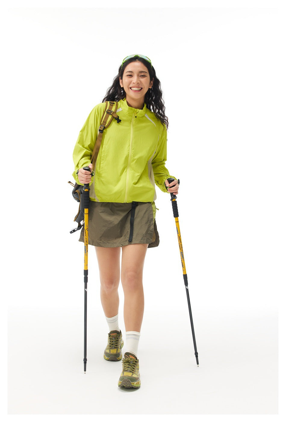 Woman in a yellow-green zip jacket and olive skirt walking with trekking poles and a backpack