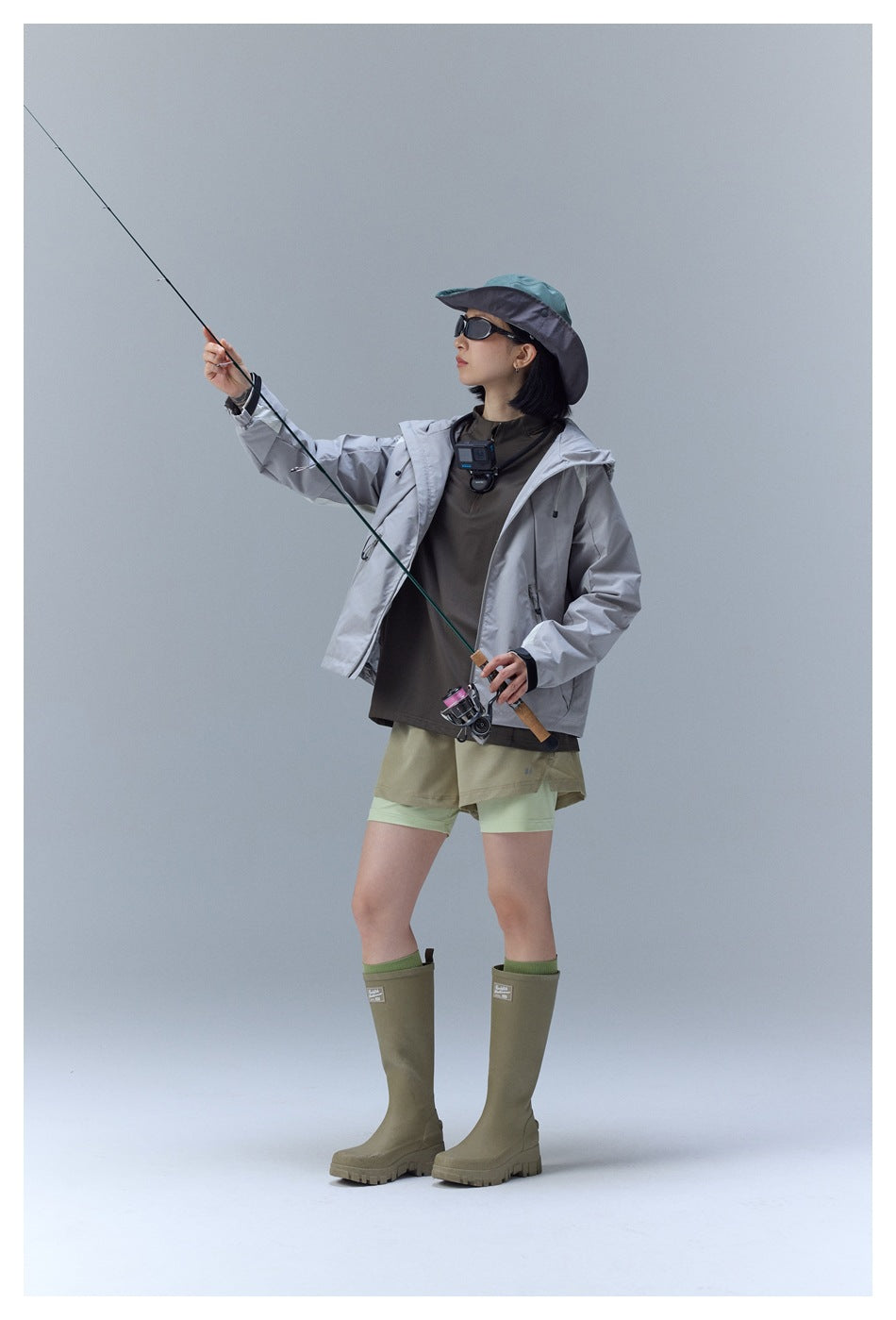 Model wearing the light gray hooded jacket open, angled pose holding a fishing rod, with shorts and tall boots