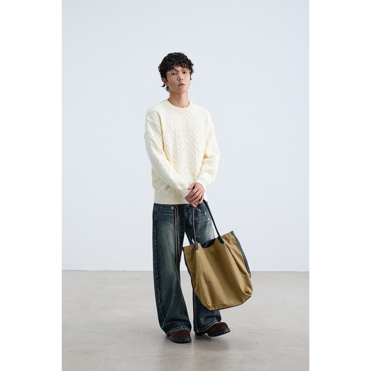 Model wearing an ivory cable-knit crewneck sweater with dark wide-leg jeans, carrying a tan tote.