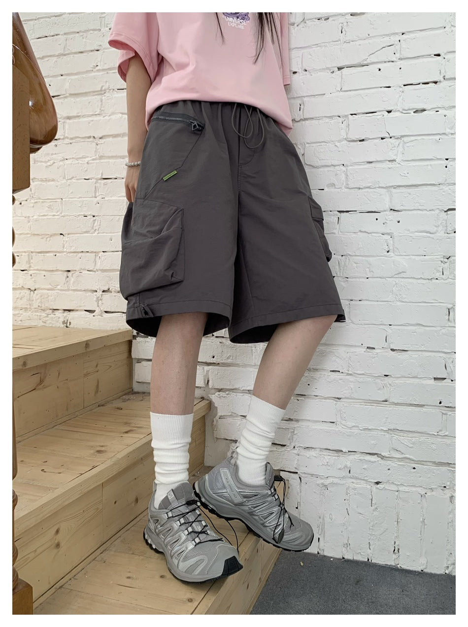 Close view of dark gray cargo shorts worn with a pink t-shirt, white socks and sneakers by a white brick wall