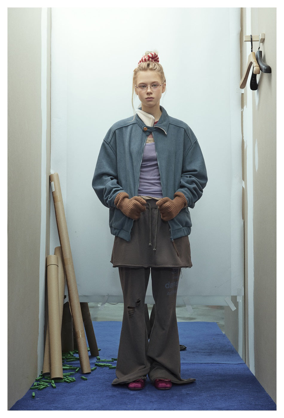 Model facing forward in a blue-gray ribbed jacket with a high collar, brown gloves, and dark layered bottoms.