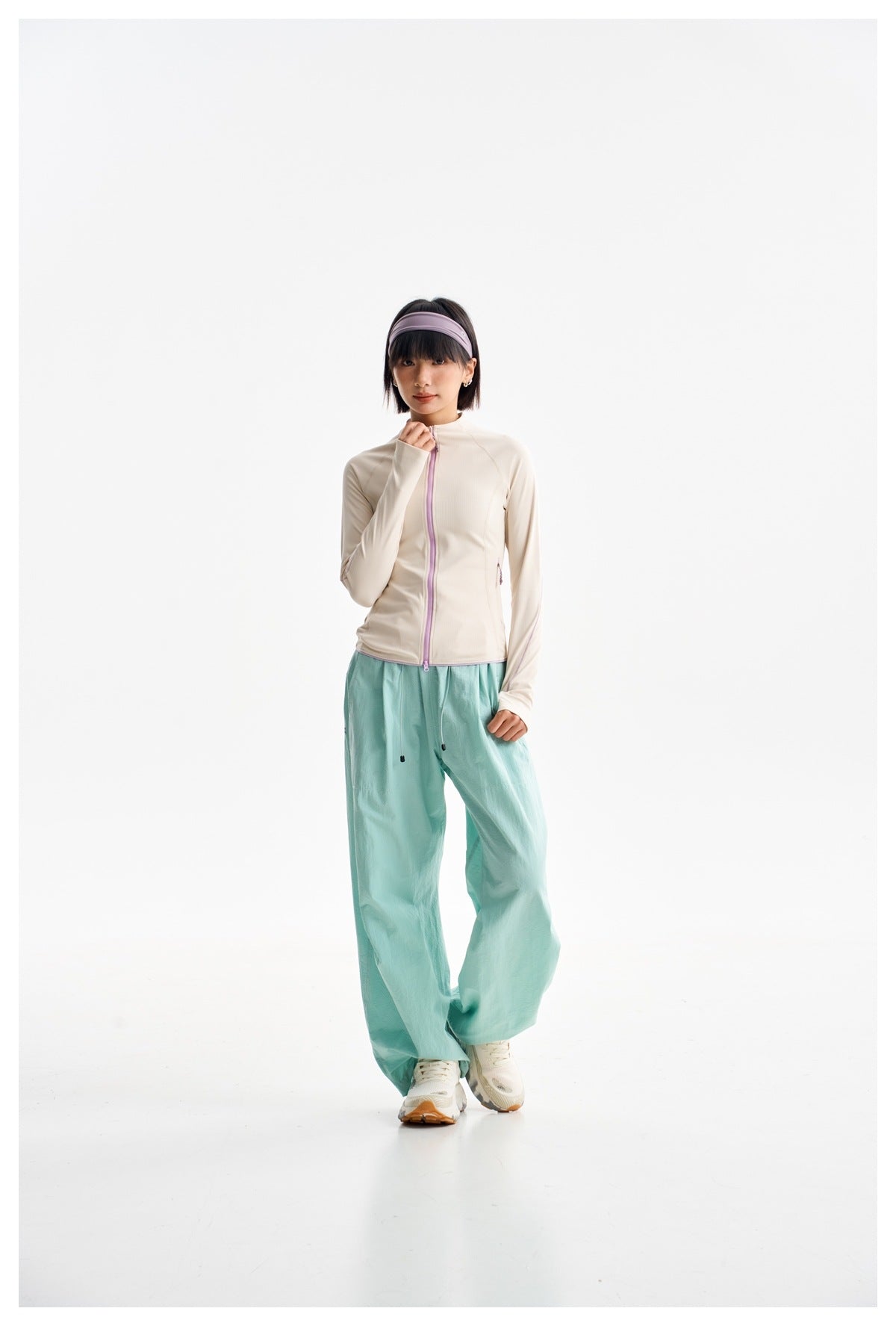 Full-length model wearing an ivory zip jacket with lilac trim, loose aqua trousers, and white sneakers.