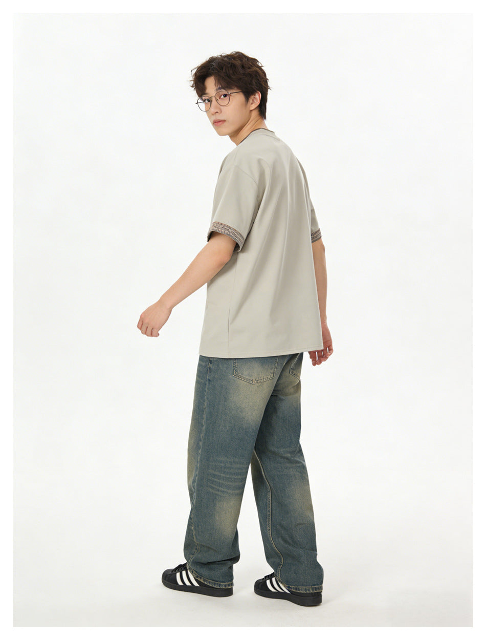 Rear three-quarter view of a model wearing a light beige T-shirt with patterned sleeve trim and loose blue jeans.