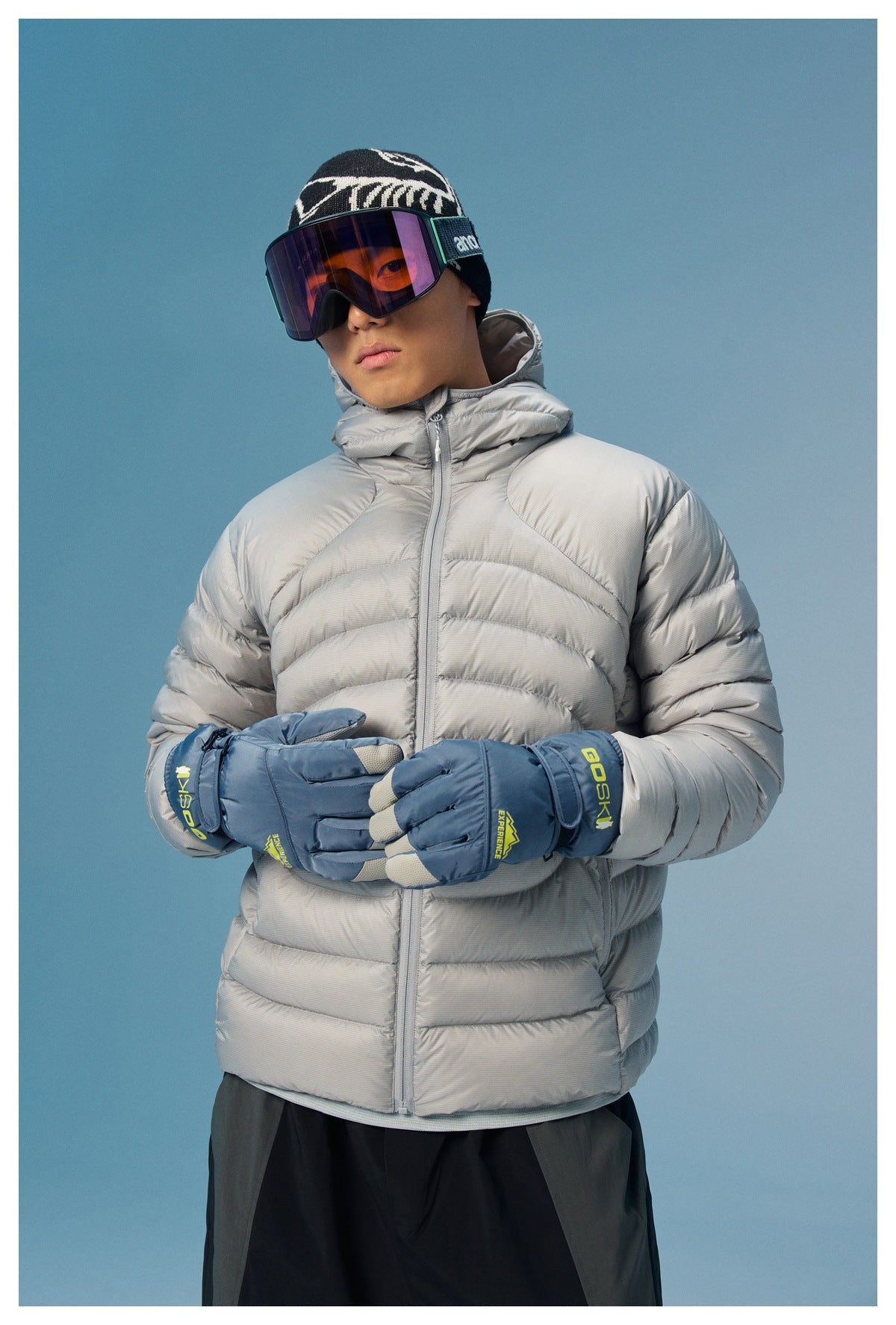 Model wearing a light gray quilted puffer jacket with a high collar and blue ski gloves, shown from the front.