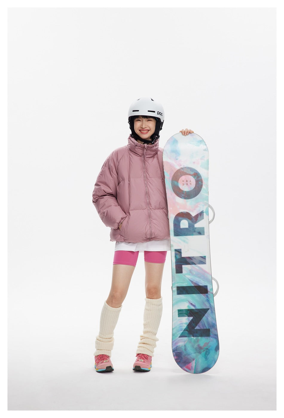 Model wears a dusty pink puffer jacket with pink shorts and holds a colorful snowboard upright.