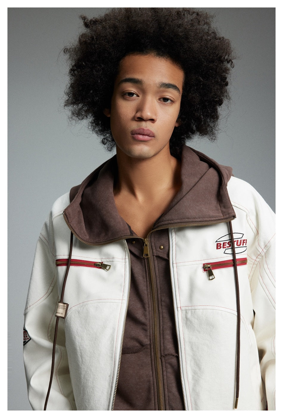 Model wearing an off-white jacket with red stitching and zip pockets over a brown hooded layer.