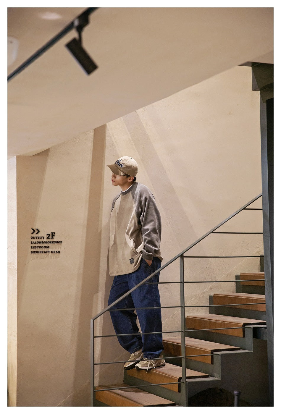 A man stands on a staircase wearing a two-tone taupe and gray raglan sweatshirt, dark jeans, and a beige cap.