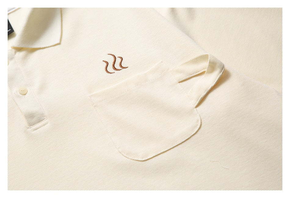 Close-up showing the ivory polo's brown embroidered chest motif, patch pocket, buttons, and textured knit.