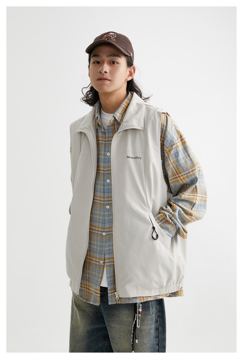Model wearing a light gray zip-front vest with drawcord pocket details over a blue and tan plaid shirt.