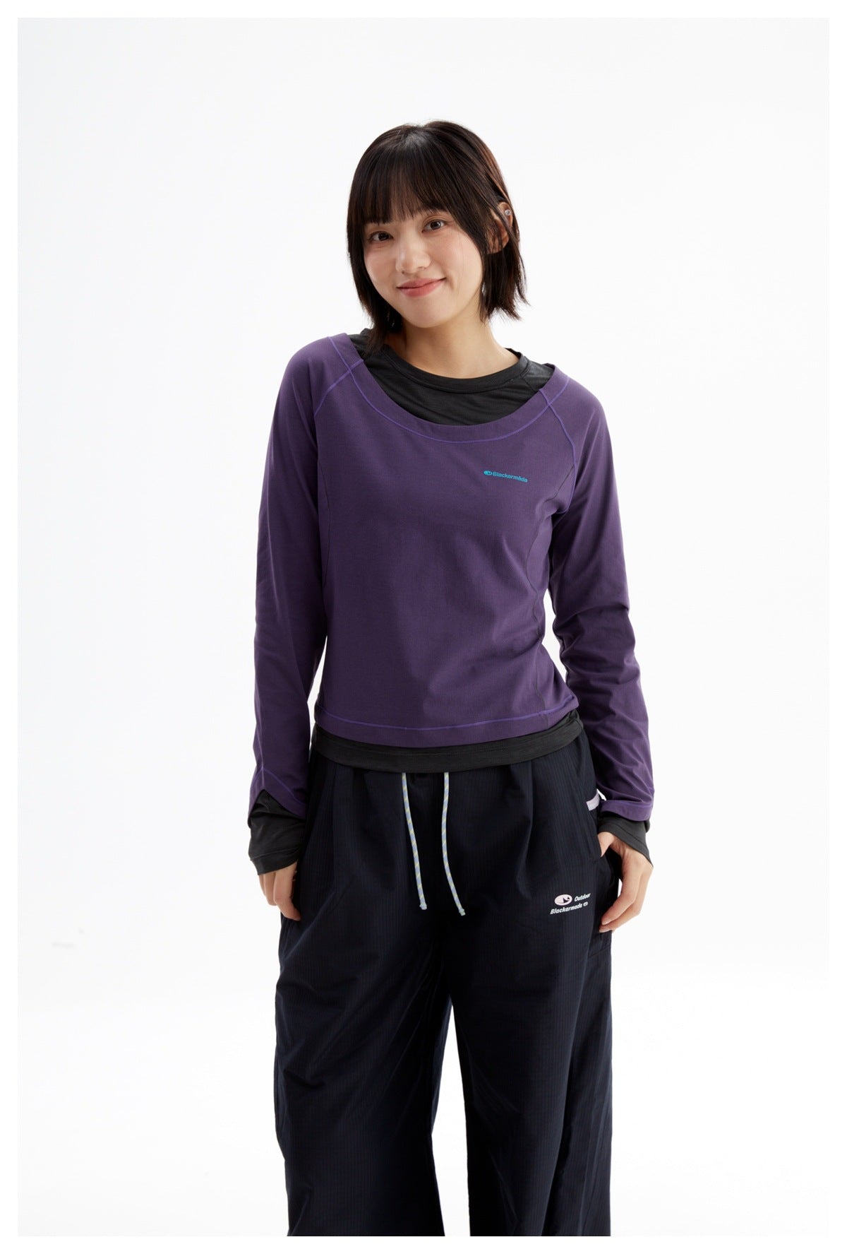 Model wearing a purple long-sleeve top with a black undershirt and loose black pants, standing at a slight angle.