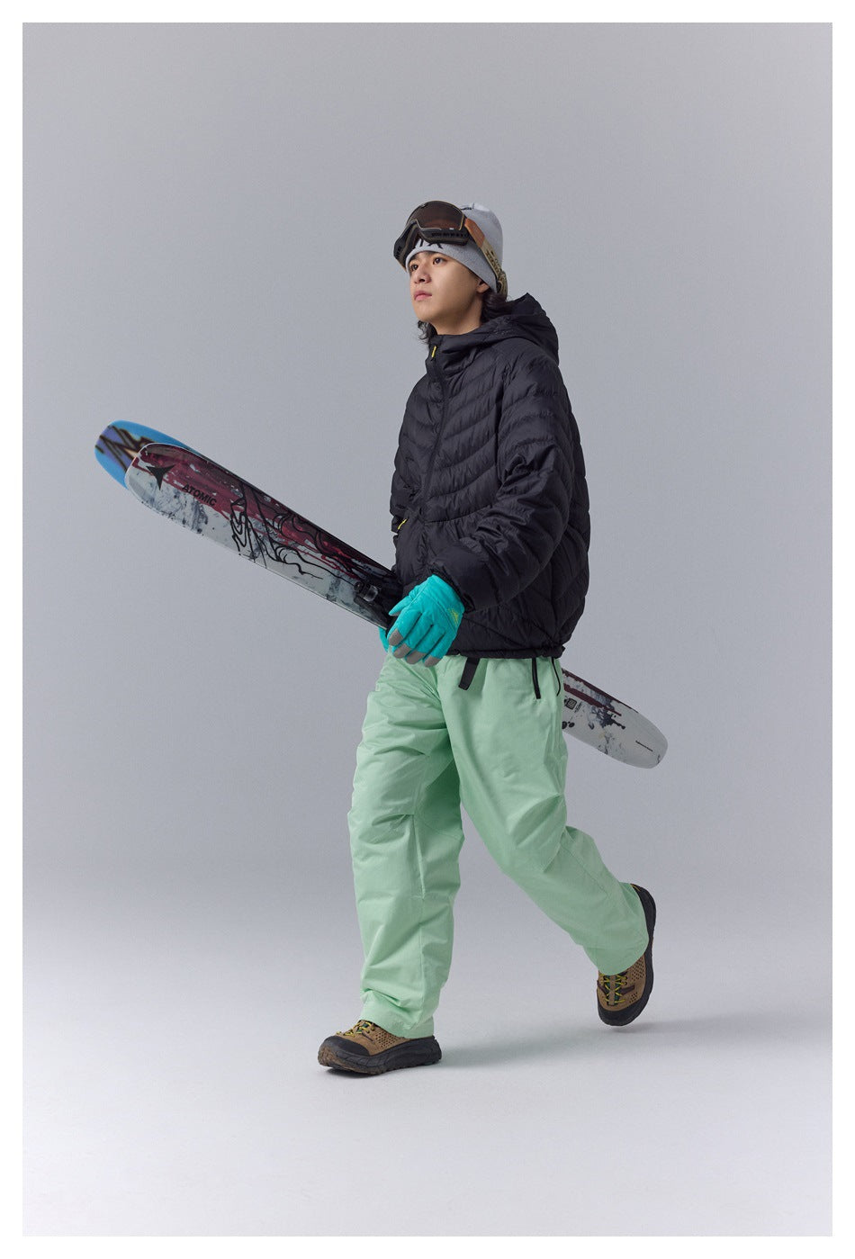 Model in a black quilted hooded jacket and pale green snow pants, carrying skis while walking.
