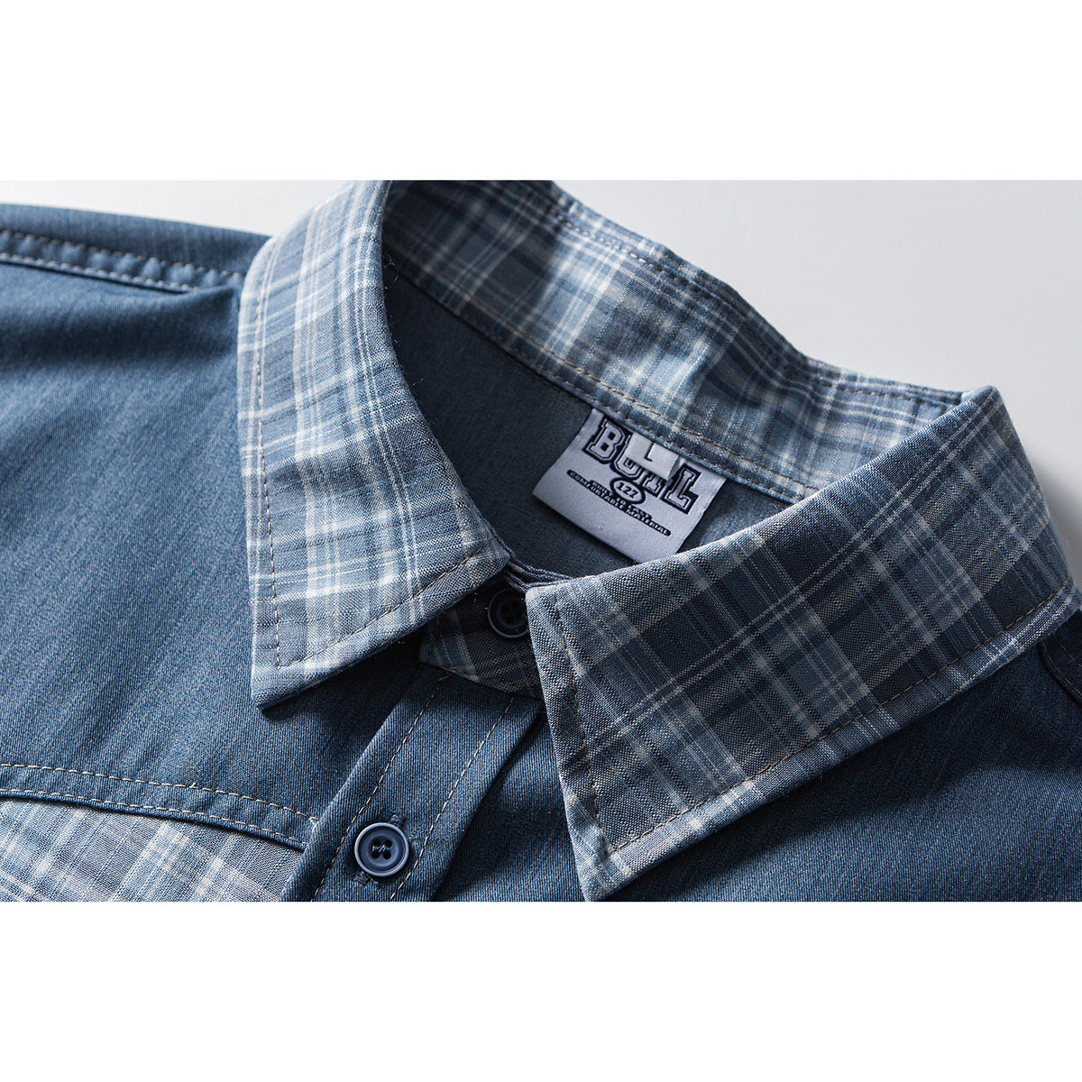 Close-up of a blue shirt collar with gray-blue plaid trim, dark buttons, and a sewn-in neck label.