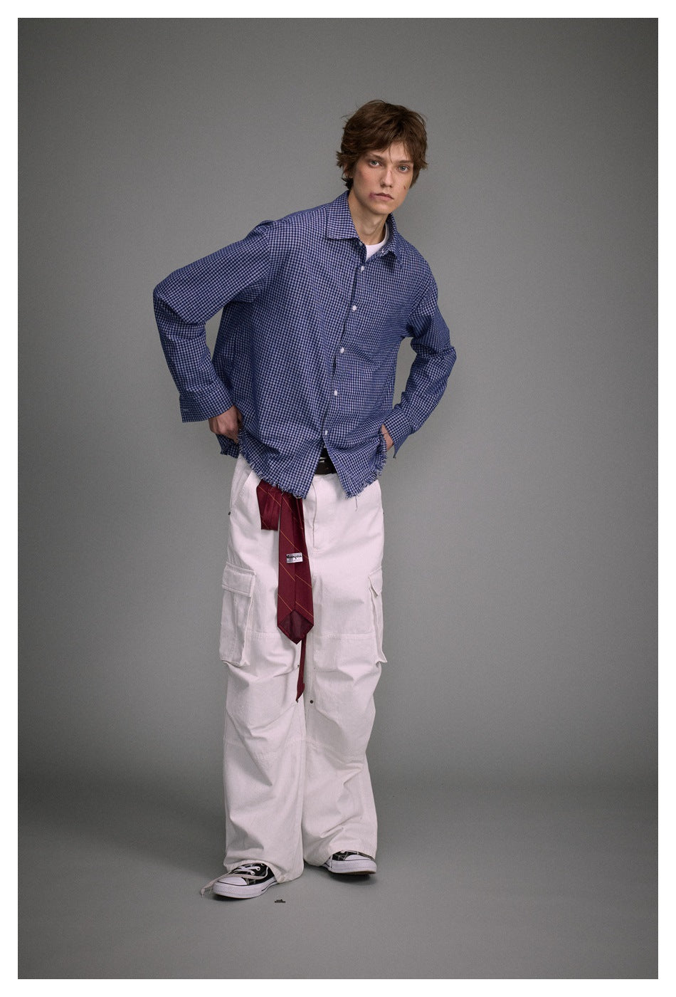 Model wearing a blue small-check button-up shirt with a frayed hem, white cargo pants, and a burgundy tie.