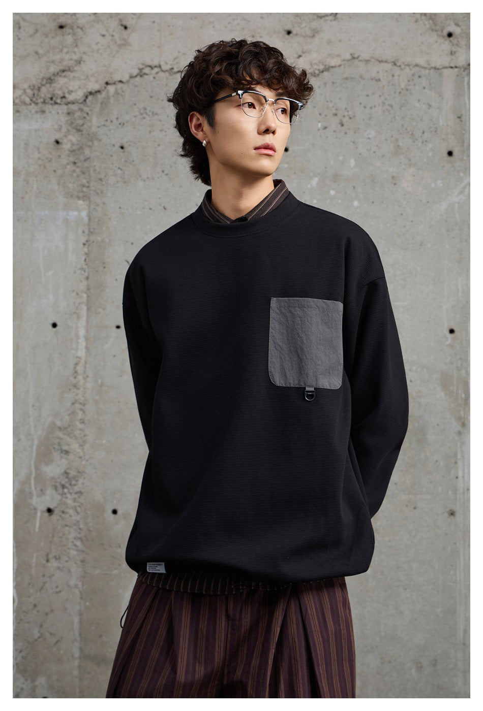 Model wearing the black sweatshirt with gray chest pocket over a striped shirt, against a concrete wall