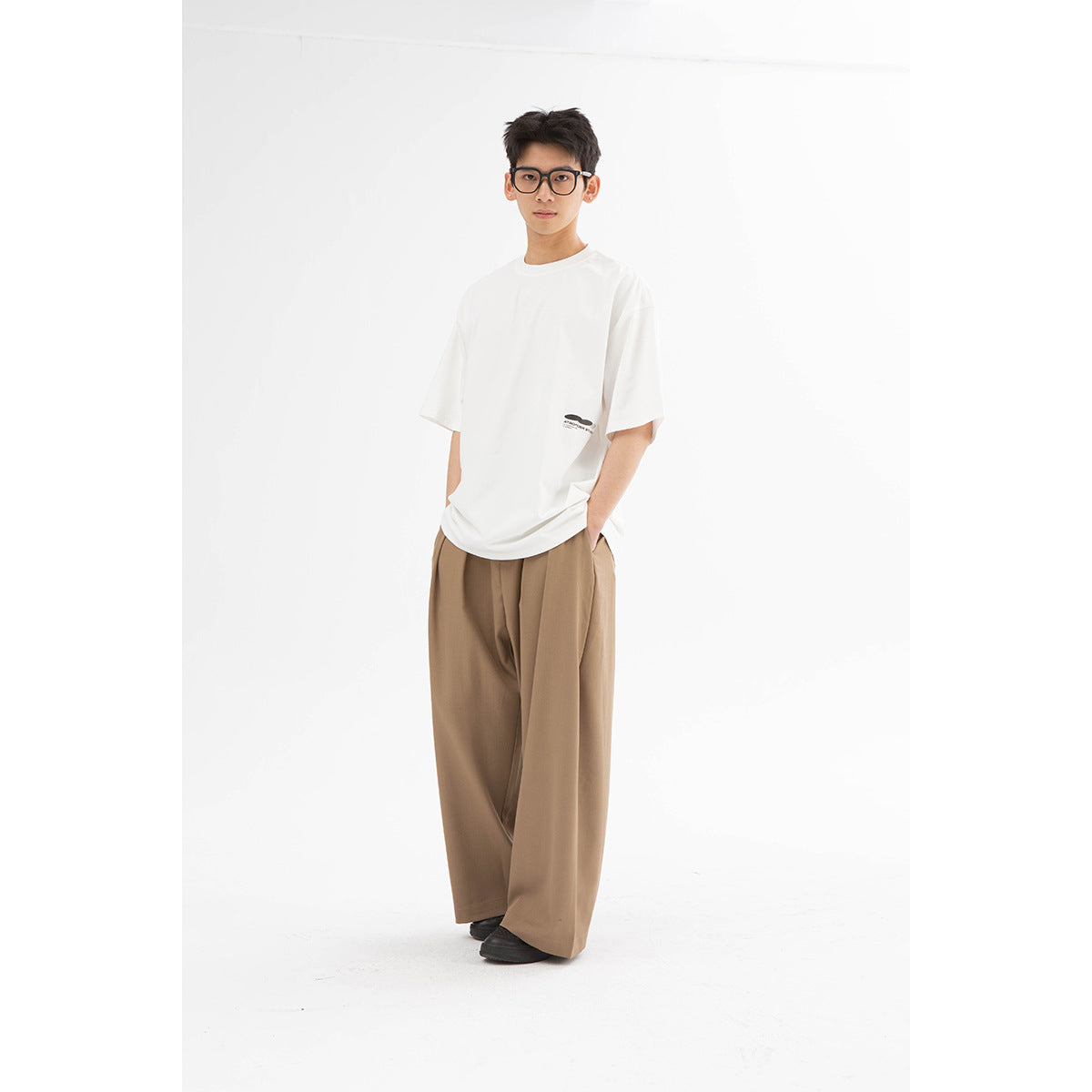 Model wearing an oversized white T-shirt with a small chest logo and wide-leg tan trousers.