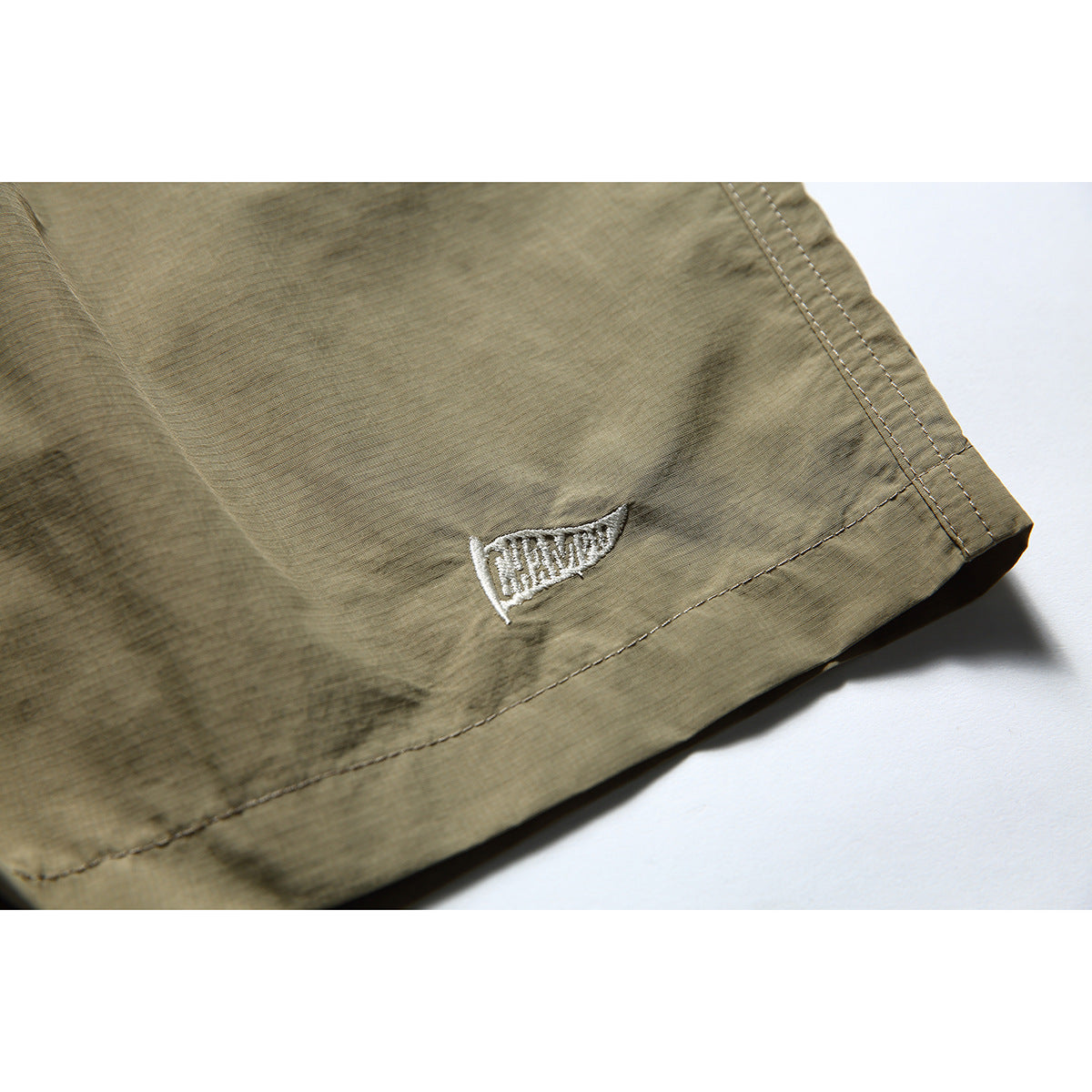 Close-up of the olive-khaki shorts' hem with a small white embroidered pennant emblem and double stitching.