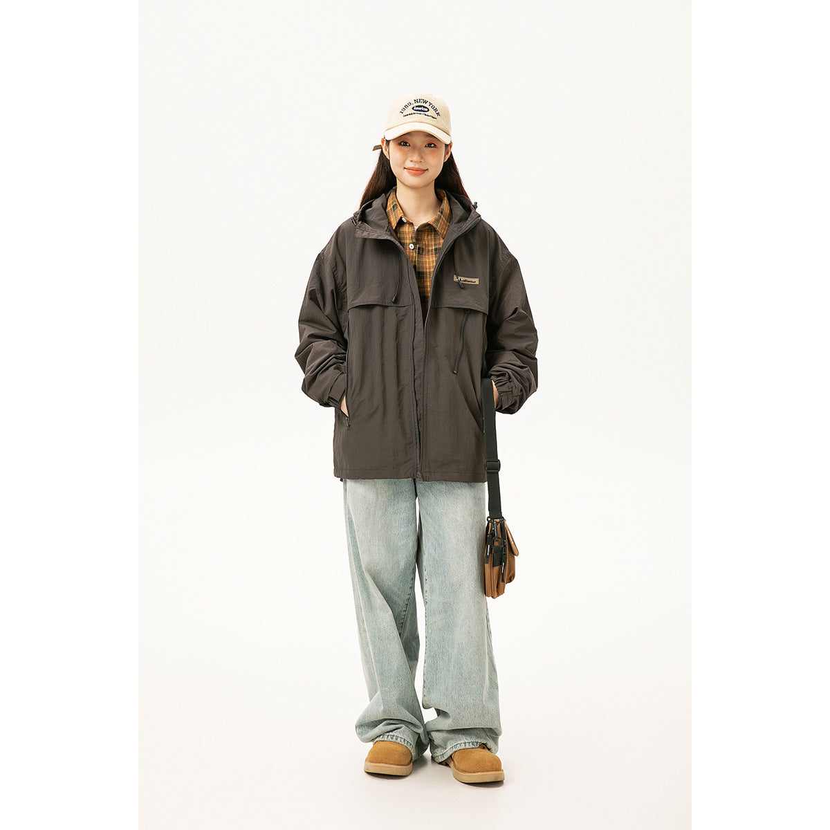 Model wearing a dark charcoal hooded jacket over a checked shirt, with loose light-wash jeans and a cap.
