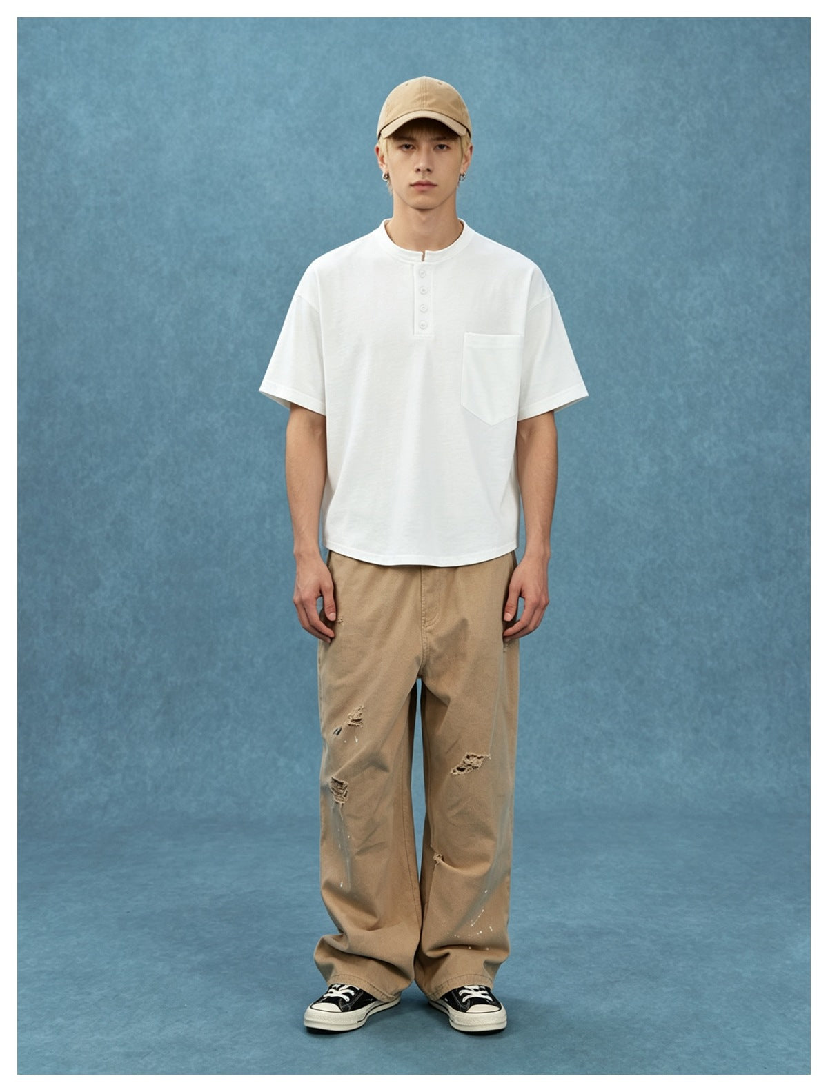 Model wearing a white Henley tee with chest pocket, beige cap and wide khaki pants against a blue backdrop
