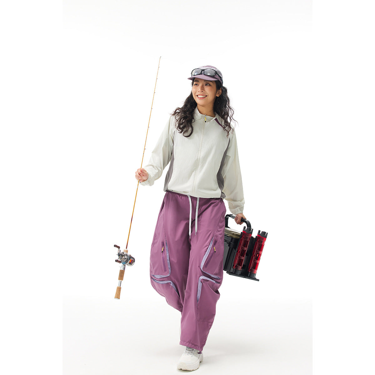 Woman wearing a white zip jacket and mauve cargo pants, holding a fishing rod and gear against a white background.