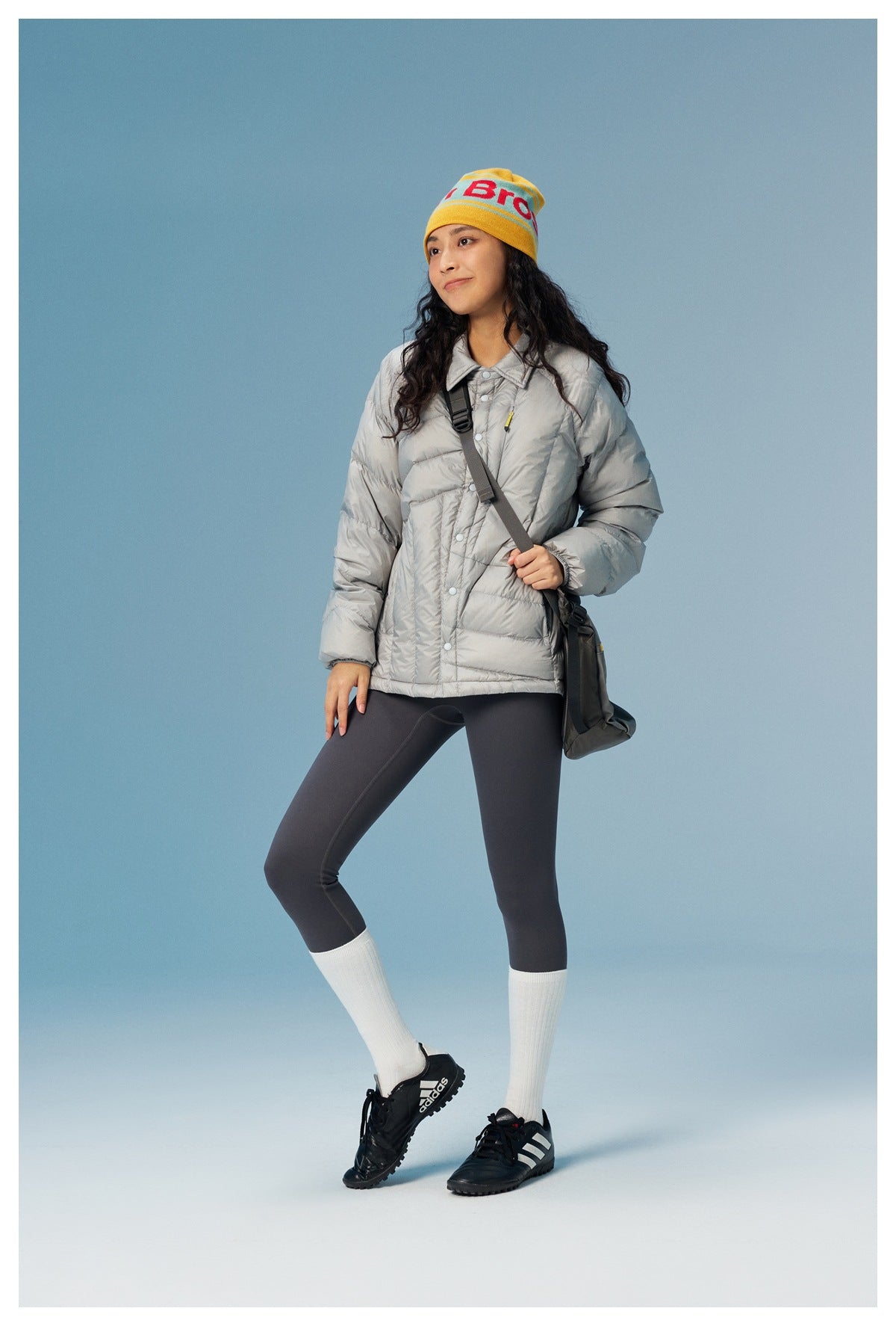 Woman wearing the gray quilted snap-front jacket with leggings, beanie, crossbody bag and sneakers against blue backdrop