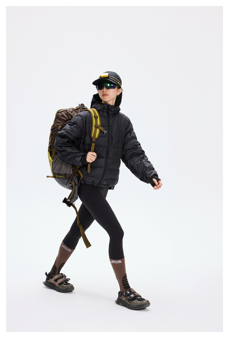 Model wearing a black hooded quilted jacket with black leggings and hiking gear, carrying a brown and olive backpack.