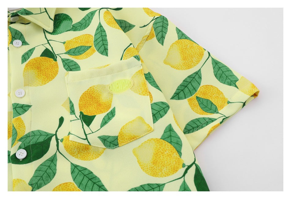 Close-up of a pale yellow button-up shirt with a lemon-and-leaf print, chest pocket, white buttons and small oval embroidery.