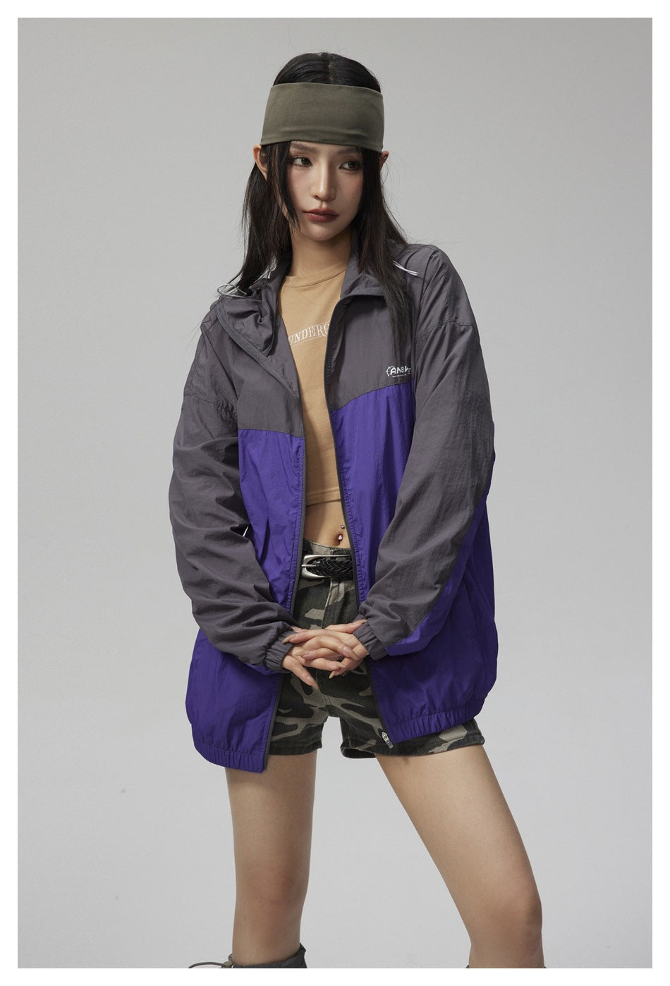 Model wearing an open charcoal and purple hooded jacket over a tan top and camouflage shorts.