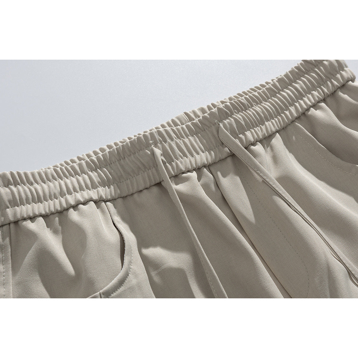 Close-up of light beige trousers showing a gathered elastic waistband, drawstring, and pocket opening.