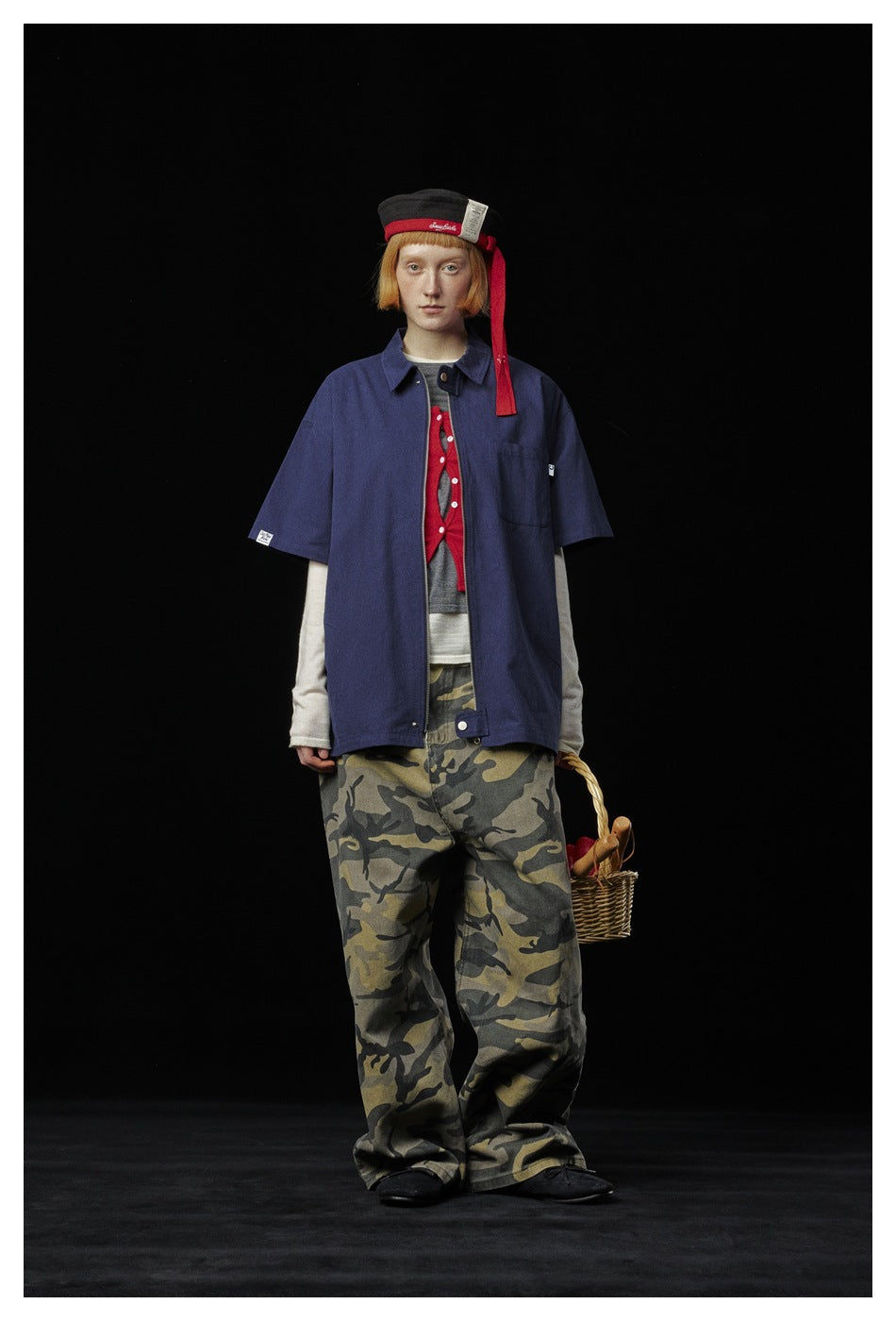 Model wearing a blue short-sleeve zip shirt open over layers with camouflage pants, front view on black background