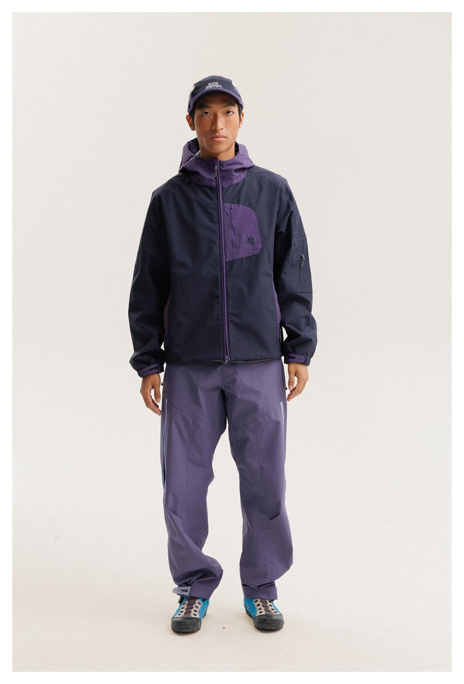 Full-length view of a model wearing a navy and purple hooded jacket with purple pants and a cap.