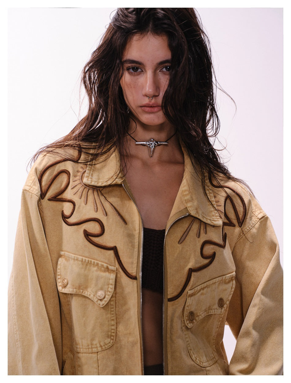 Close view of a model wearing an oversized tan jacket with dark embroidered motifs, a zip front, and flap pockets.
