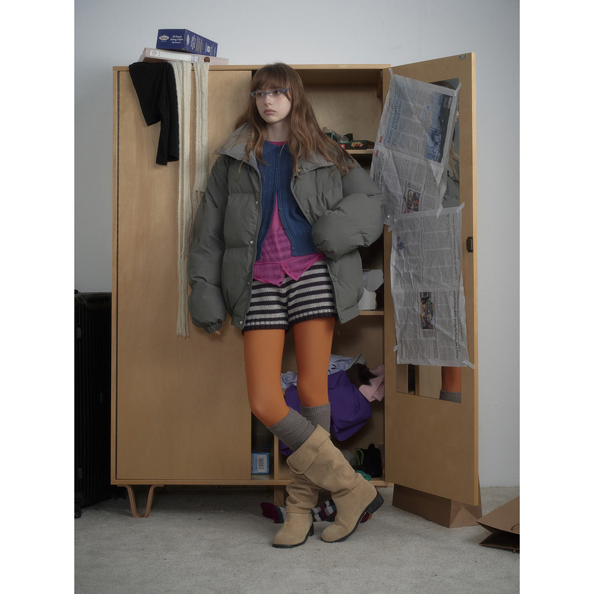 Model wearing an open charcoal-gray puffer jacket with a wide collar, layered over colorful knitwear and striped shorts.