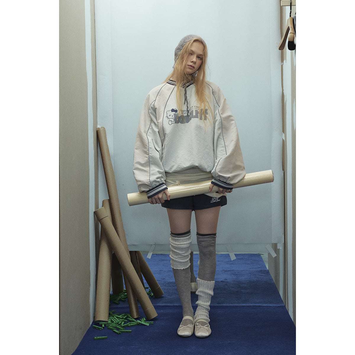 Model wearing an oversized cream pullover with gray panels, striped trim, and a cartoon text graphic, with black shorts.