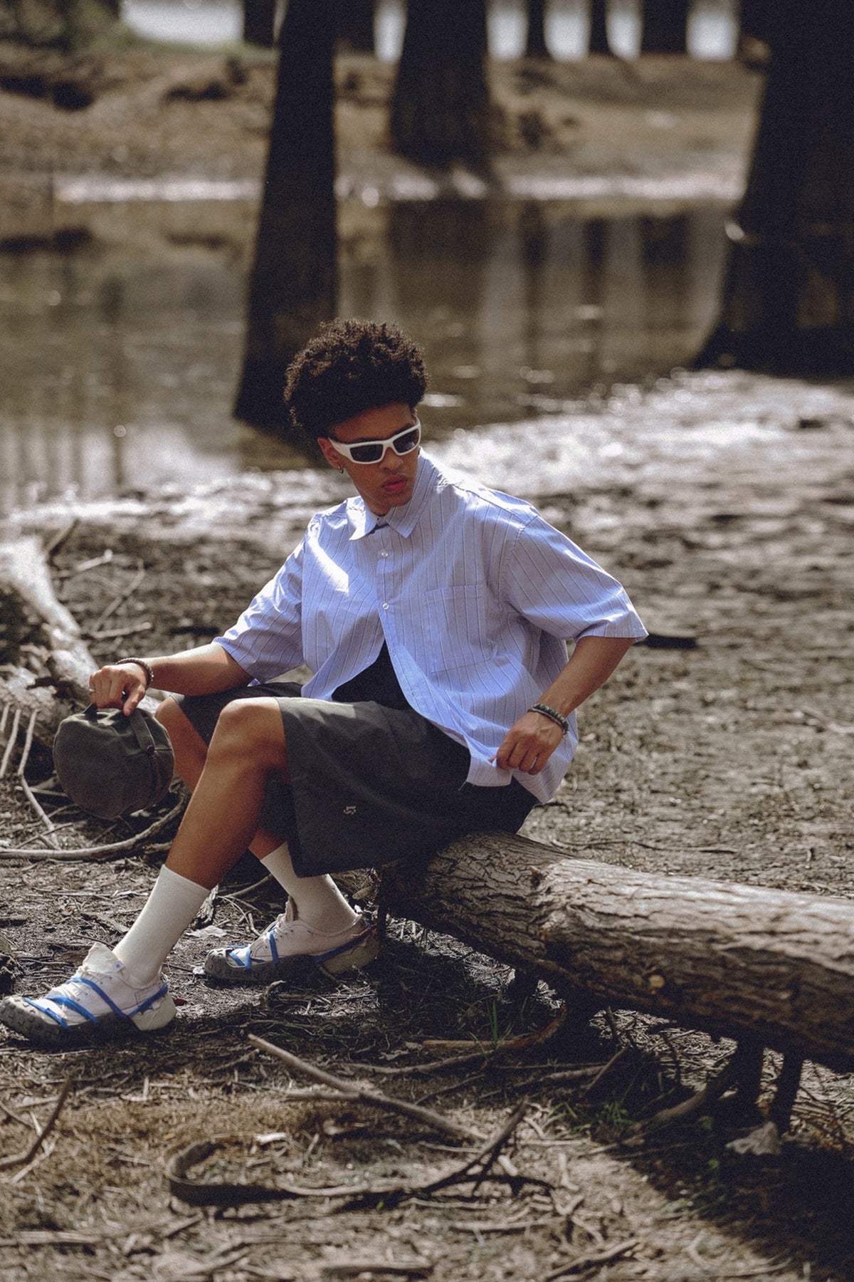 Seated model wearing a pale blue pinstriped short-sleeve shirt and dark shorts outdoors.