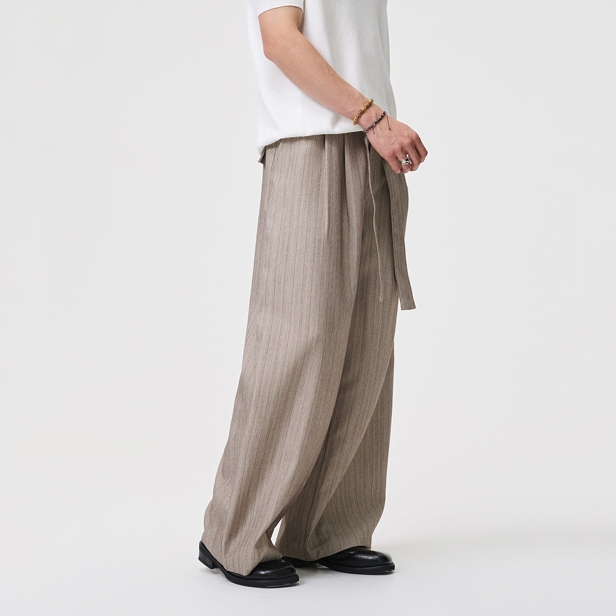 Side view of a model wearing loose taupe-gray pinstriped trousers with a long waist tie and black shoes.