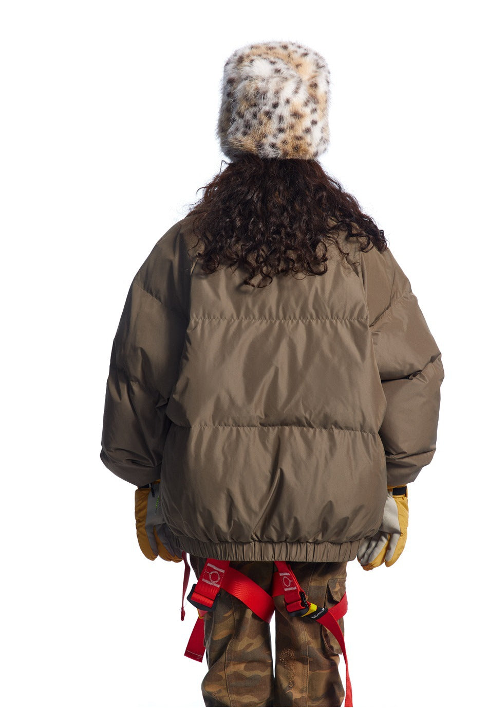 Back view of a dark taupe oversized puffer jacket worn with camouflage trousers and a spotted fur hat.