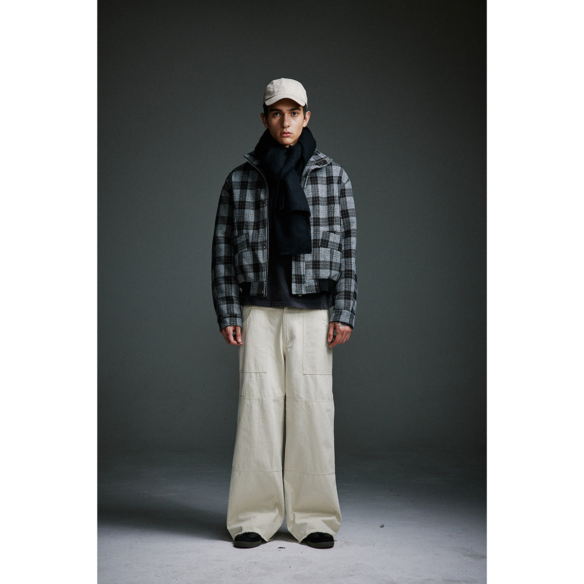 Full-length model wearing a gray-and-black plaid zip jacket, black scarf, cream wide-leg trousers, and a light cap.