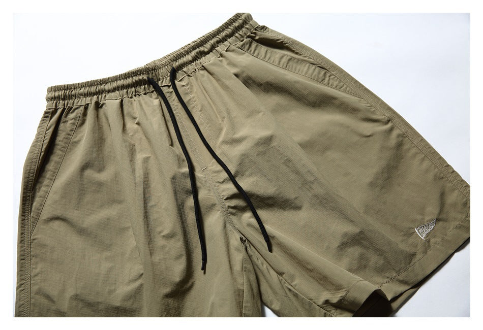 Angled close-up of olive-khaki shorts showing the elastic waist, black drawstrings, side pocket and small hem emblem.