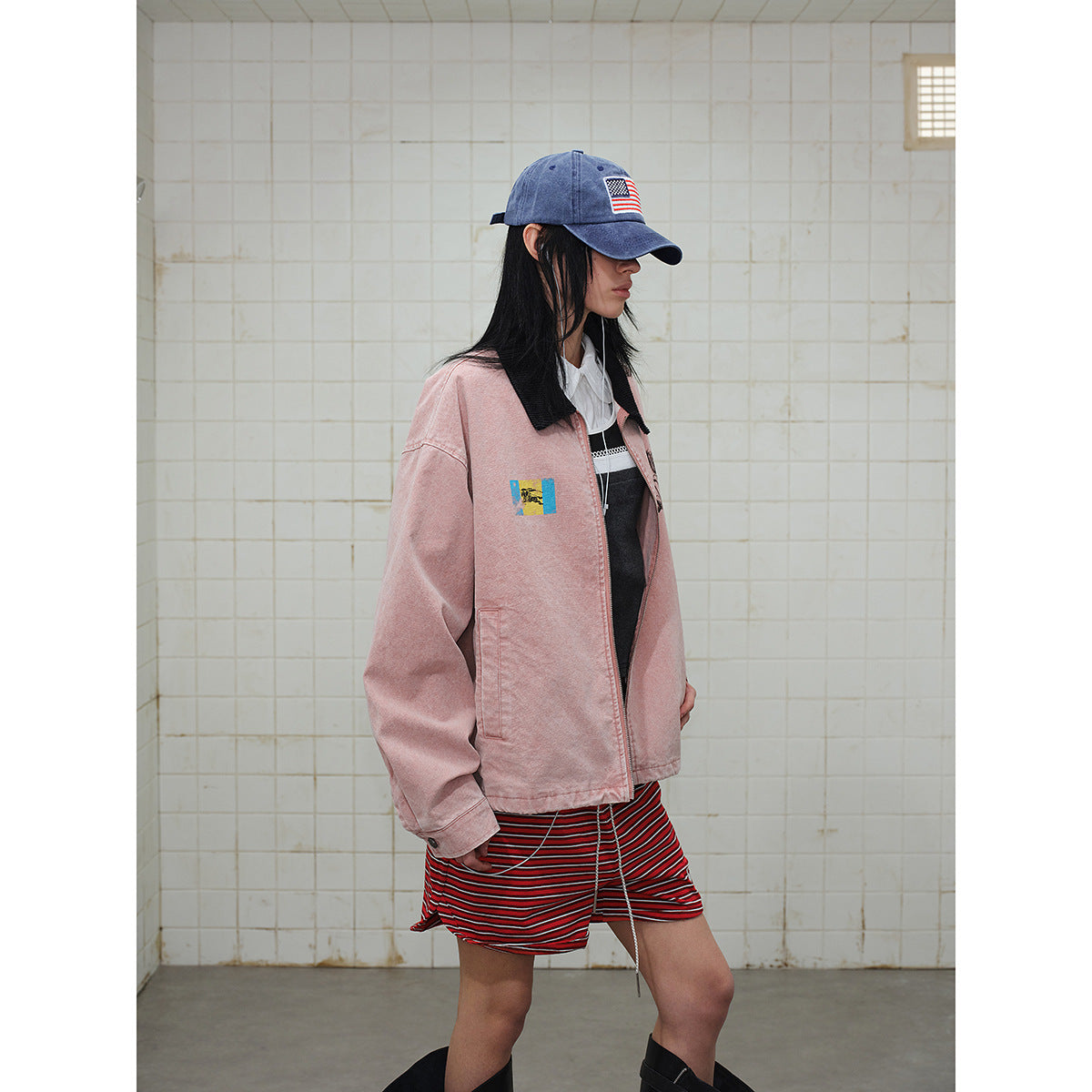 Model in a dusty pink jacket with a black collar and graphic patches, red striped shorts, and a blue cap.