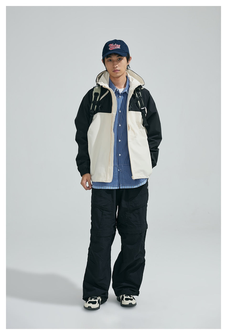 Model wearing black and beige hooded jacket over a blue checked shirt with black cargo pants, front view