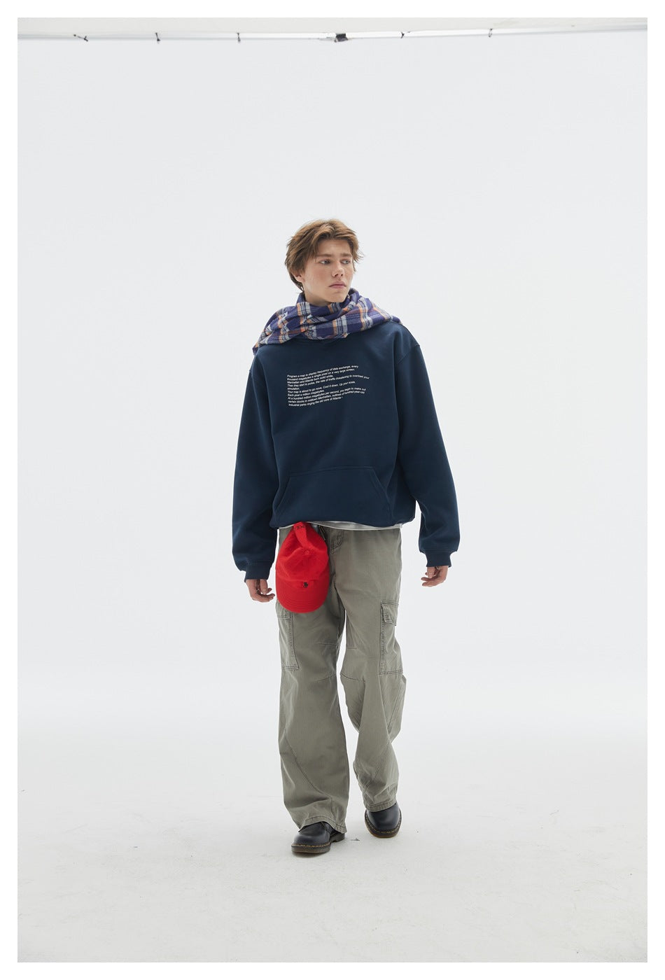 Model wearing a navy crewneck sweatshirt with small white text, plaid scarf, and olive cargo pants.