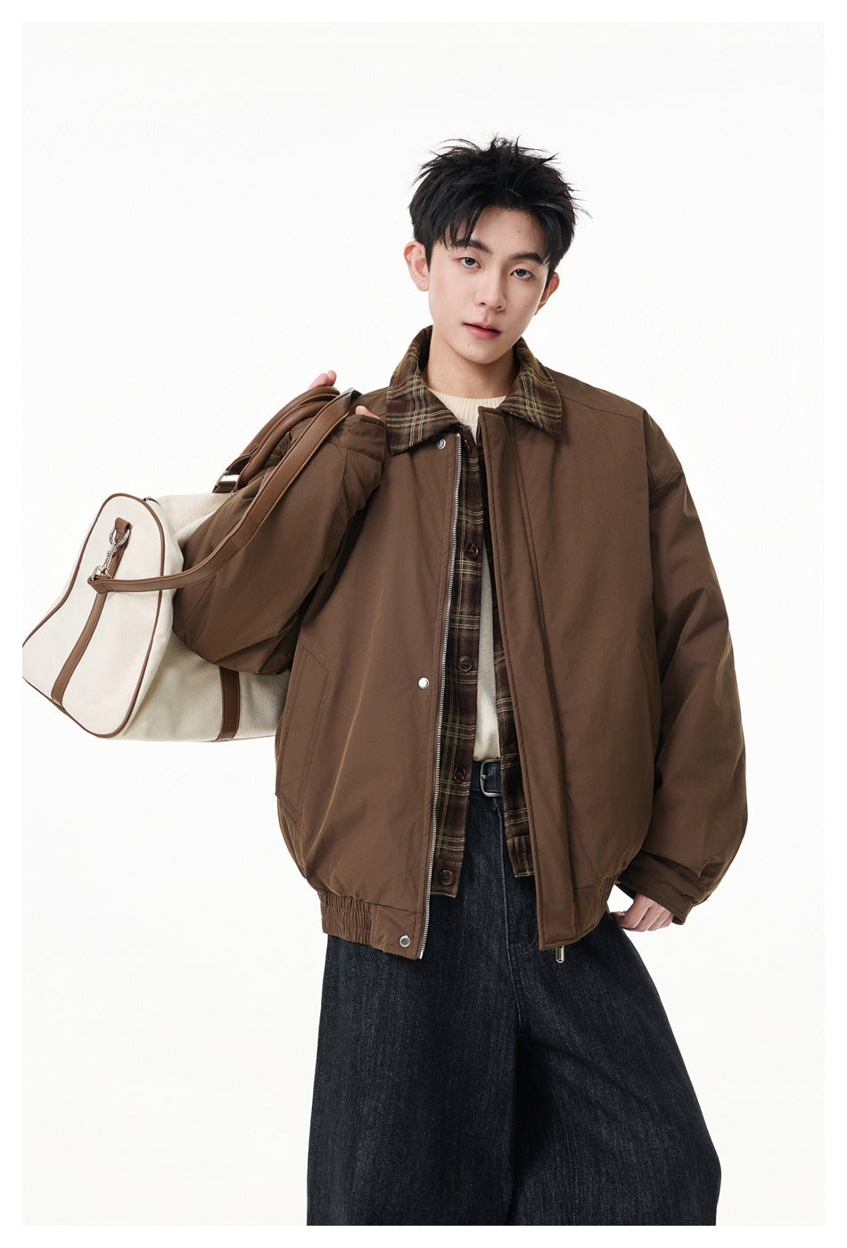 Model in a brown jacket with plaid collar and trim, carrying a cream duffel bag over one shoulder.