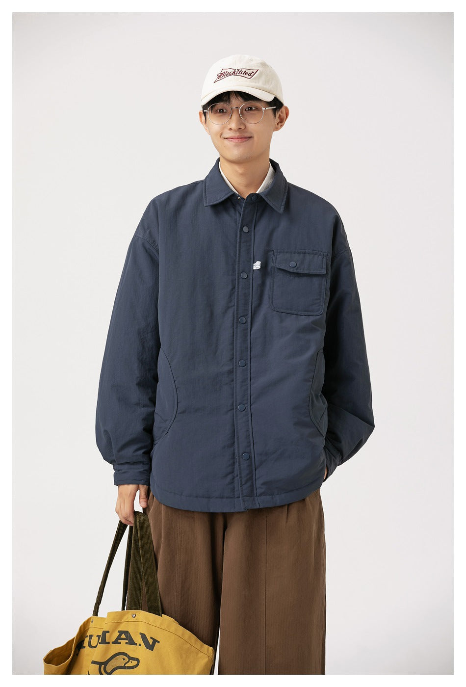 Model wearing a navy snap-front jacket with chest flap pocket, cap and brown pants, carrying a tote bag