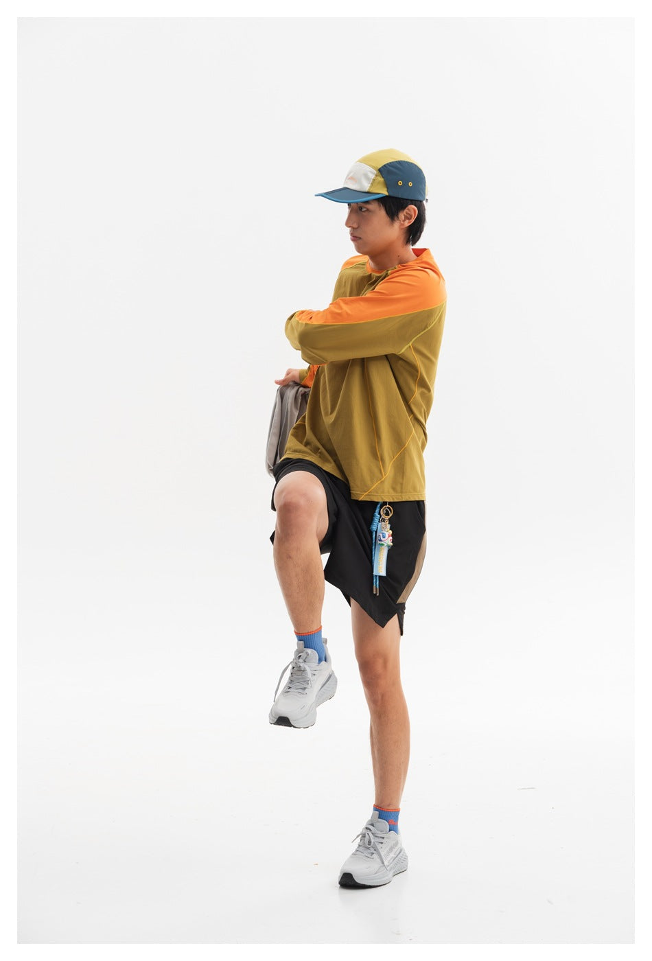 Side view of a model wearing an olive and orange long-sleeve top with black shorts and a color-block cap.