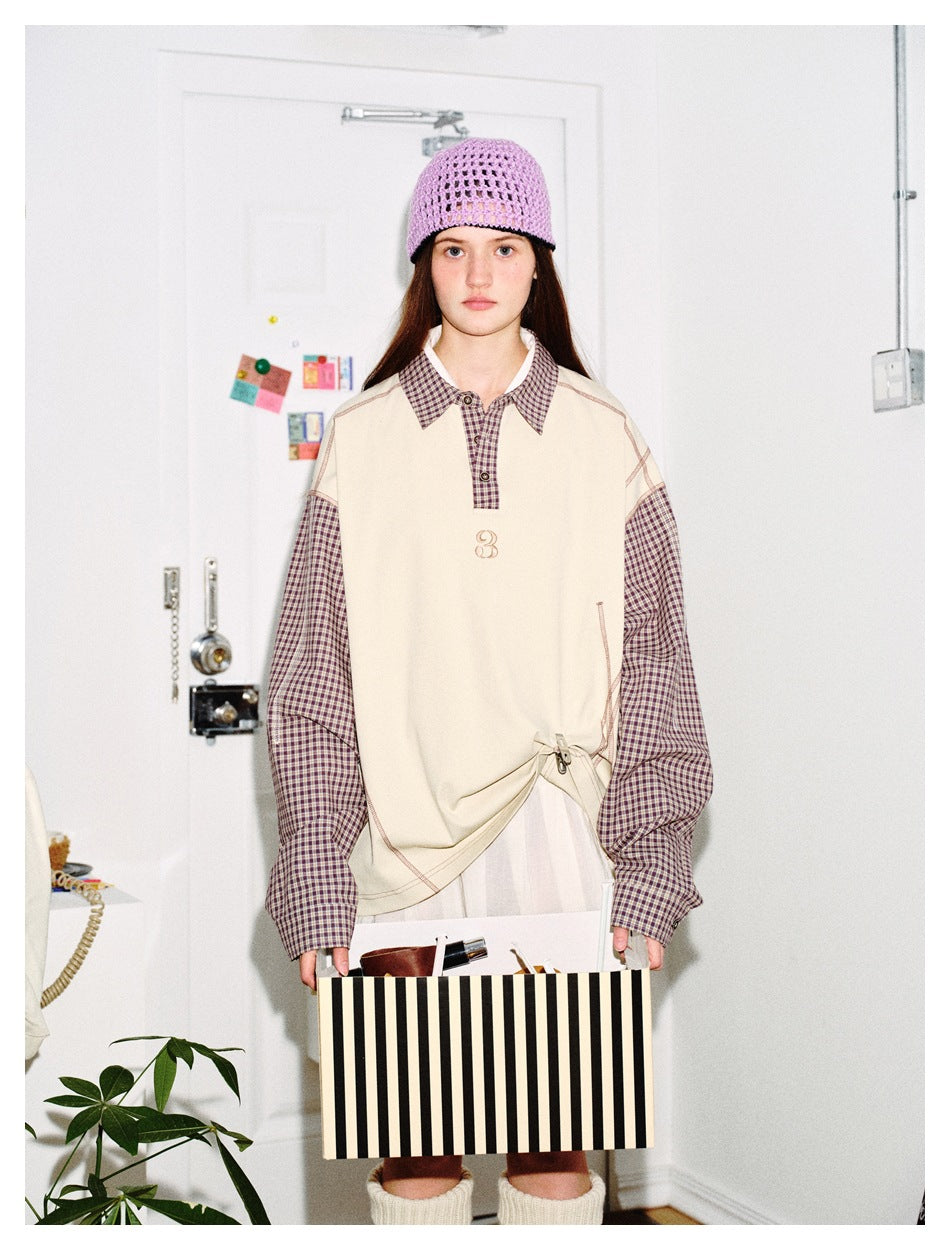 Model wearing an oversized cream top with brown plaid sleeves and collar, a lilac knit hat, and a white pleated skirt.