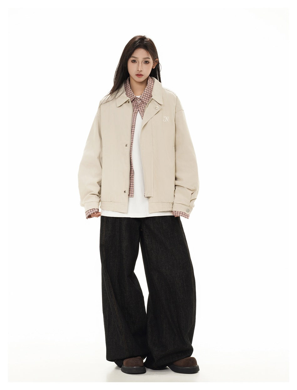 Full-length model wearing a light beige collared jacket over a pink checked shirt and white top, with loose black jeans.