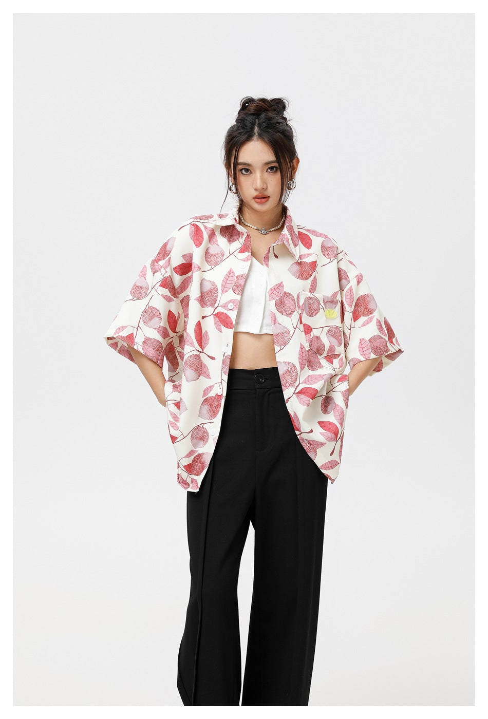 Model wearing a white short-sleeve button-up shirt with pink leaf print, styled open over a white crop top and black