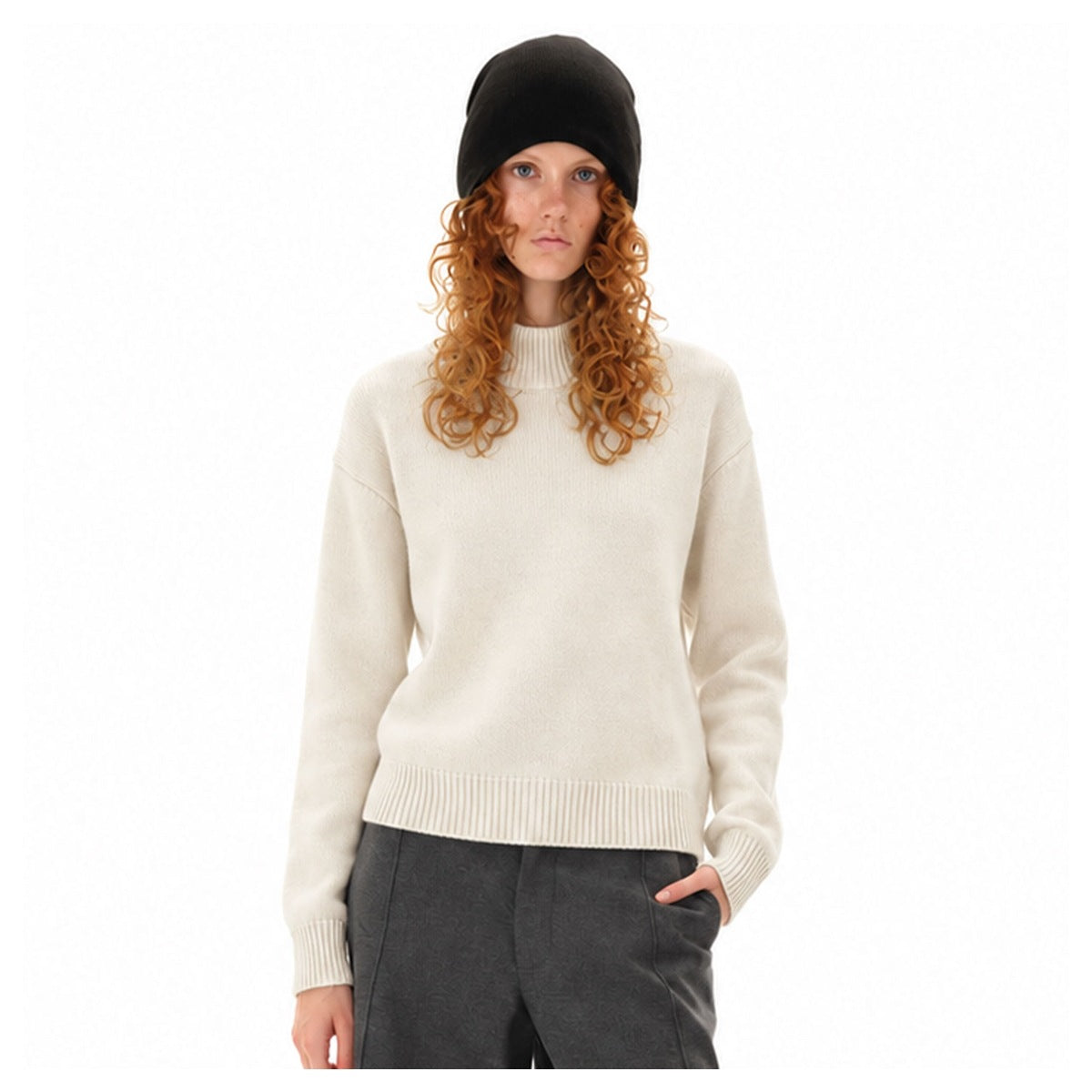 Woman wearing an ivory high-neck sweater with ribbed trim, dark trousers and a black beanie.