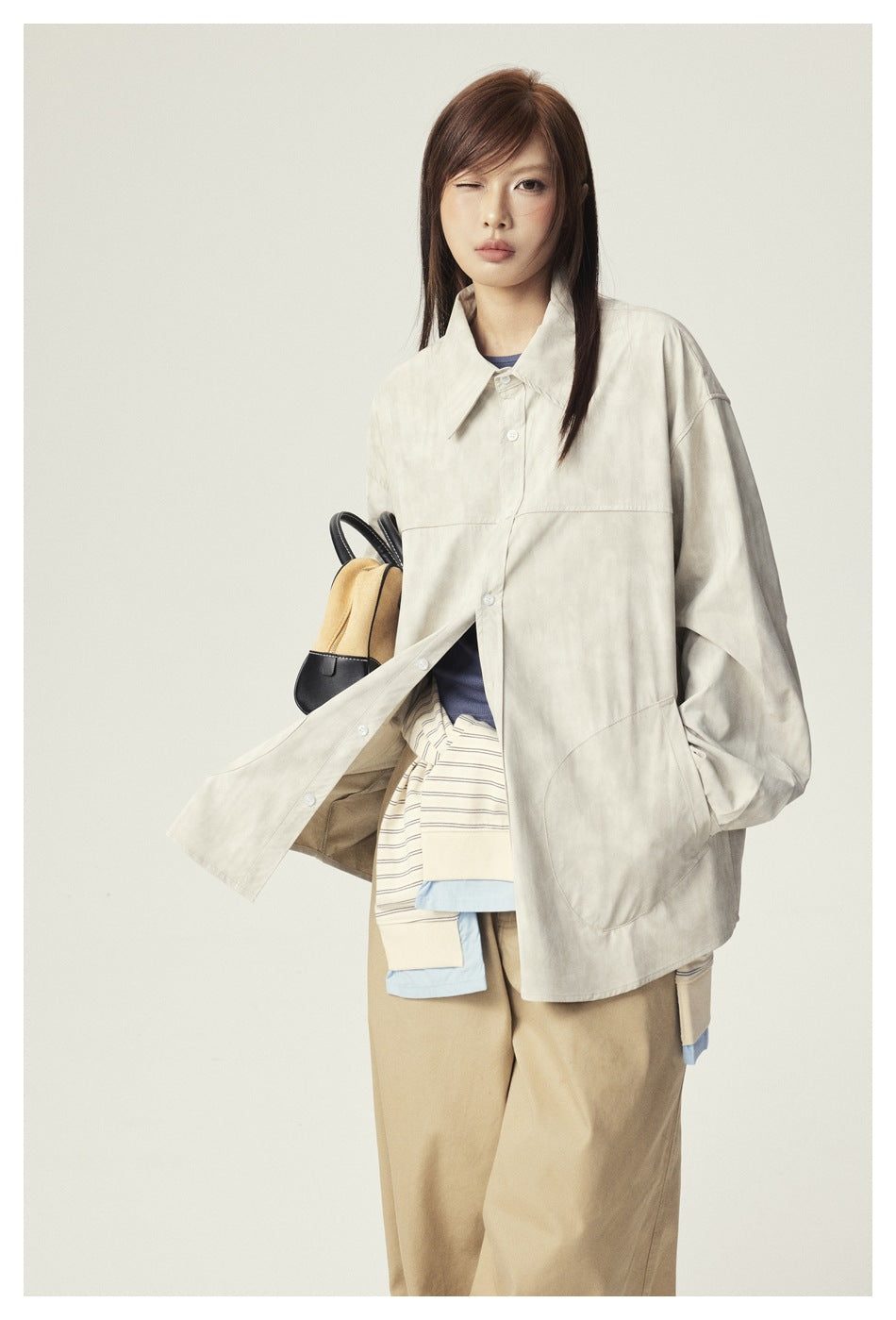Model wearing an oversized ivory button-up jacket with large patch pockets, layered over a striped top and tan trousers.